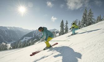 Two skiers are skiing in the snowy mountains. The sun is shining and the sky is clear.