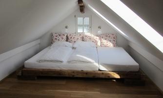 A cozy bedroom in the attic with a large bed and floral pillow. Large windows let in plenty of daylight.