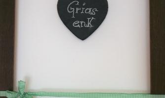 A brown wooden frame with a slate heart on which "Griaß enk" is written. The frame is decorated with a green check ribbon.