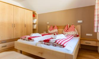 A cozy bedroom with a double bed and red-and-white checked pillows. Wooden cabinets and a light wooden floor give the room a warm atmosphere.