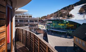 A view from a balcony overlooking a busy street and the ski resorts in the background. On the opposite side stands a modern building with a large display.