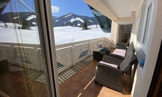 A balcony with seating and a view of snow-covered mountains. The sun is shining and the sky is clear.