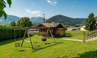 A beautiful garden with a playground that includes swings and a tire slide. In the background, green mountains and a blue sky can be seen.
