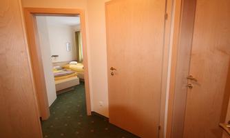 A hallway with wooden doors and green carpet. In the background, a room with two beds and a window is visible.