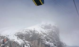 A gondola floats over snow-covered mountains beneath a cloudy sky. The landscape appears wintry and impressive.