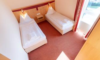 A simple room with two single beds and carpeted flooring. The room walls are light, and there is a large window with daylight.