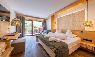 A modern hotel room with wooden details and a large window. There is a double bed, comfortable furniture, and a small balcony with a view.