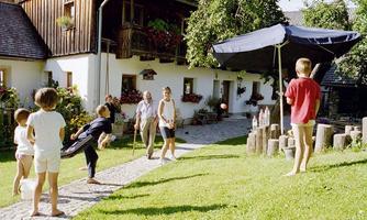 A group of children is playing in the garden in front of a traditional house. An older person is observing the scene, while a sun umbrella stands in the background.