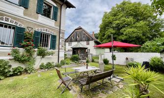 A beautiful garden with a seating area and a red sun umbrella. In the background, there is a charming old house with green shutters.