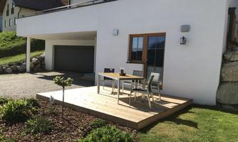 A modern house with a wooden terrace and an outdoor table. The garden is well-maintained and there is a garage.