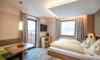 A modern room with a double bed, a table, and a seating area. Large windows provide ample daylight and offer a view outside.