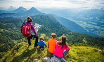 A family is sitting on a hill and looking at a map. In the background, green valleys and mountains stretch under a clear sky.