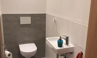 A modern guest bathroom with gray and white tiles. There is a sink, a toilet, and a towel holder with a red towel.