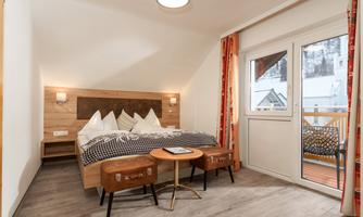 A modern bedroom with a large bed, wooden details, and a balcony. Bright curtains and cozy furniture create an inviting atmosphere.