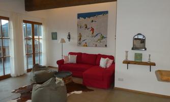 A cozy living room with a red sofa and comfortable cushions. A large painting hangs on the wall and large windows provide plenty of daylight.