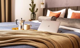 A cozy bedroom with a bed, a laptop, and a coffee pot on the blanket. On the nightstand, there is a small plant and a cup.