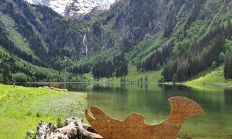 A picturesque lake surrounded by green hills and high mountains. In the foreground, there is a wooden sculpture of a fish.