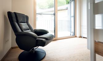 A modern black recliner is situated in a bright room. Large windows provide plenty of daylight and a view outside.