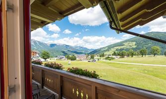 A beautiful view from a balcony overlooking a green landscape with mountains in the background. The sky is clear and blue with a few clouds.