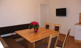 A modern dining area with a large wooden table and six chairs. In the background, there is a small kitchenette and a television on the wall.