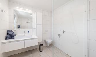 A modern bathroom with a bright, minimalist decor. There is a sink, a shower, and towels on the shelf.