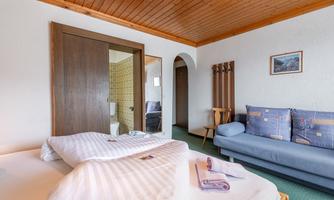 A cozy hotel room with a double bed and a sofa. The walls are light, and the wooden ceiling adds a warm atmosphere.