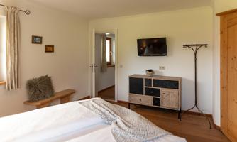 A cozy bedroom with a large bed and wood-colored furnishings. A television hangs on the wall, and there are several windows that provide natural light. The door leads directly into the living room Chiara.