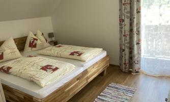 A cozy bedroom with a double bed and decorative pillows. Large windows with a view outside provide plenty of light.