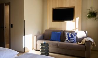 A modern living room with a beige sofa, a blue cushion, and a television on the wall. The room is designed to be bright and friendly.