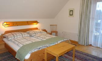 A cozy bedroom with a double bed and warm wooden furniture. Large windows let in plenty of light and provide access to a balcony.