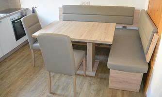 A modern dining area with a wooden table and beige seating. The kitchen is visible in the background and the floor is made of light wood.