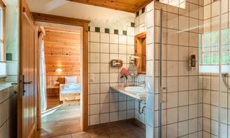 A modern bathroom with tiles and a shower. In the background, a door leads to a cozy bedroom.