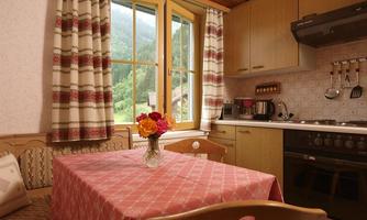 A cozy kitchen corner with a pink tablecloth and a vase with flowers. The room has large windows overlooking nature and is furnished with wooden shelves and appliances.