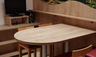 A cozy dining area with a round wooden table and high chairs. The walls are decorated with a landscape painting and wooden elements.