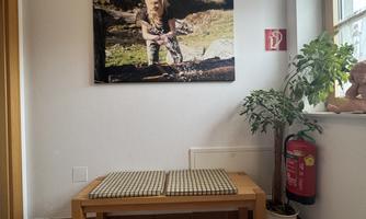 A cozy room with a wooden table and two cushions. A picture of a girl in nature hangs on the wall, and a plant is standing in the window.