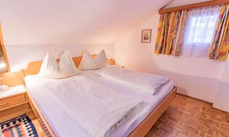 A cozy bedroom with a large double bed and two pillows. The walls are bright, and the windows are adorned with colorful curtains.
