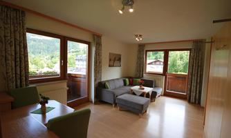 A bright living room with large windows and a view of nature. It is modernly furnished with a gray sofa and a dining table.