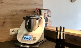 A modern kitchen robot stands on a dark work surface. In the background, a cookbook and a wall clock are visible.