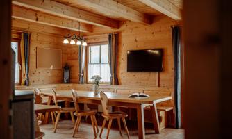 A cozy dining area in a wooden house with a large table and chairs. Large windows let in a lot of light, creating a relaxed atmosphere.