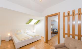 A bright bedroom with a large bed and a wooden floor. In the background, a living room with a cozy seating area is visible.