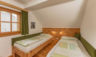 A cozy bedroom with two single beds and wooden walls. Natural light streams through the window and there are curtains in a soft green.
