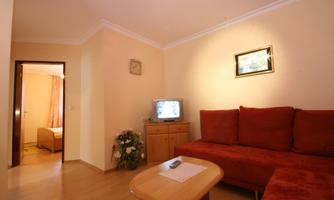 A cozy living room with a red sofa and a television. In the background, a room with a bed and curtains is visible.