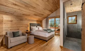 A modern bedroom with wooden paneling and large windows. There is a comfortable bed, an armchair, and an adjoining bathroom.