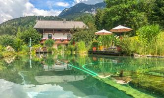 A quiet garden with a clear pond and a beautiful view of the mountains. In the background, there is a house surrounded by trees and plants.