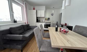 A modern living room with a cozy gray sofa and a wooden dining table. The kitchen is open and well-equipped, with light walls and large windows.
