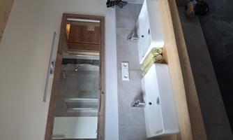 A modern bathroom with two sinks and a large mirror. The décor is simple and elegant, featuring wood and stone elements.