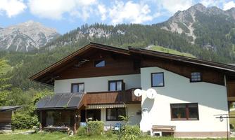 A beautiful house in the mountains with a large green meadow. In the background, tall mountains and blue sky can be seen.