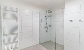 A modern bathroom with a walk-in glass shower. The walls are covered in white tiles, and a towel radiator can be seen.