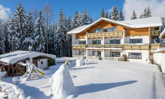 A beautiful chalet in the snow, surrounded by tall, snowy trees. The sky is clear and blue, and the property is covered with a white blanket of snow.