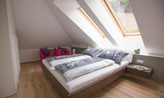 A modern bedroom with a double bed and slanted windows. The decor is bright and cozy, with a small seating area in the corner.